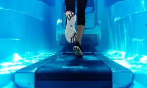 Underwater view of a person's legs and feet on a treadmill, performing aqua jogging in a bright blue pool.