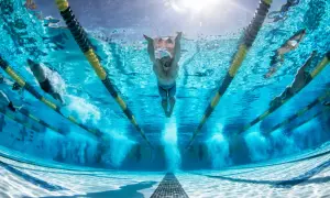 Underwater view of swimmers in a pool, with a focus on a swimmer performing butterfly stroke. The sun shines through the water, illuminating the lanes and the powerful stretches for swimmers.
