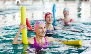 Three smiling fitness swimmers in a pool, holding yellow pool noodles.