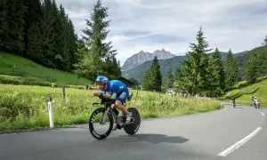 Athlete in blue cycling gear races on a road bike during a sprint triathlon, with a scenic mountain backdrop.