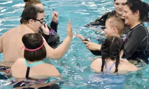 A group enjoys swim lessons for special needs, with instructors and participants high-fiving and smiling in the pool.