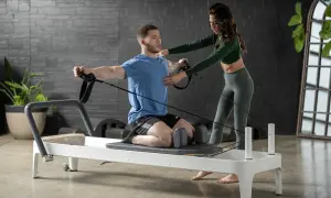 A man on a reformer uses resistance bands for pilates for swimmers, with a trainer guiding his form.
