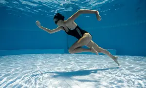 Underwater shot of a woman in a black swimsuit performing aqua aerobics. Her body is streamlined as she propels herself through the blue water.