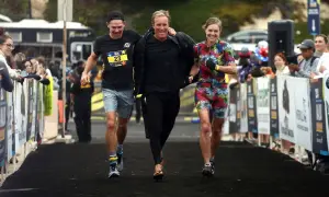 Three friends celebrate crossing the finish line of an Ironman relay. The woman in the colorful floral suit beams, while the two men, one in a black shirt with a race bib, share a laugh.