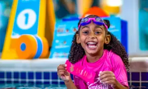 A happy girl in goggles and a pink rash guard holds a Goldfish Swim School ribbon. She's in a pool, ready for swim classes for 5 year olds.