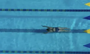 A swimmer glides through the water, executing a powerful breaststroke. The blue pool is marked with lane lines and colorful flags.