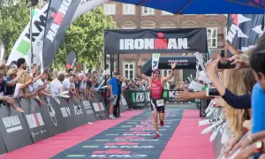 A triumphant athlete crosses the finish line at an Ironman triathlon, arms raised in victory, cheered by a crowd.