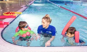 A smiling swim instructor in a blue shirt teaches two young boys in a pool. They are using pink pool noodles for support.