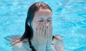 A young woman in a pool covers her nose and mouth, eyes closed, possibly experiencing a chlorine allergy sneezing fit.