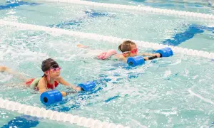 Two girls with goggles swim in a pool, holding blue flotation devices. One girl's face is partially submerged, showing her determination.
