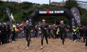 Athletes in wetsuits run from the water under an "Ironman Swim" arch. The start of their race is a sandy beach, ready for the iron man swim challenge.