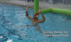 A water polo goalie in a red cap with hands raised, ready to block a shot.