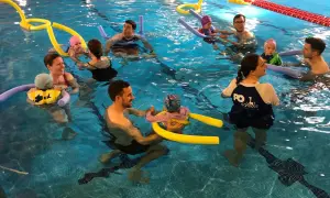 Parents and babies enjoy mommy and me swim classes, splashing and playing with pool noodles in the bright blue water.