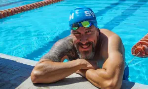 A smiling man in a blue swim cap and goggles rests on the pool edge, ready to teach how to swim laps for beginners.