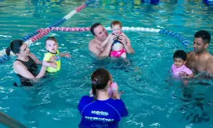 Parents and babies enjoy a swim tots class in a bright blue pool. An instructor in a blue shirt with "Aqua-Tots" visible helps a baby.