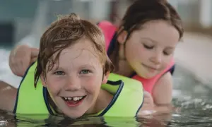 A happy boy with a missing tooth grins during 3 year old swim lessons, with a girl in a pink life vest behind him.