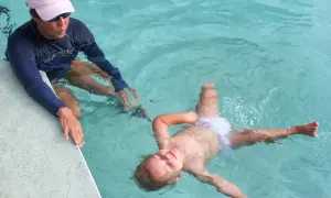 A child floats on their back in a pool, learning to swim. An instructor in a blue rash guard and pink hat watches.