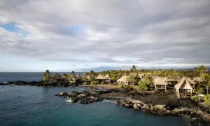 Tropical bungalows on a rocky coast in Kona, Hawaii. The calm ocean reflects the cloudy sky, a stark contrast to the power of a tsunami.