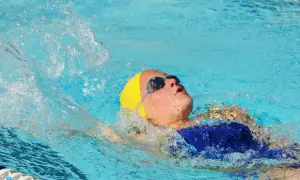 A young swimmer with a yellow cap and goggles performs the back stroke, creating a splash as they move through the blue water.