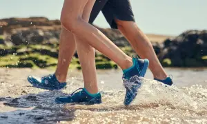 Two people wearing aqua jog shoes splash through shallow water on a rocky beach.