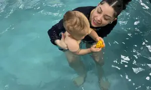 A woman holds a baby in a pool, both smiling. The baby plays with a yellow toy. This scene shows that you can swim with tubes in your ears.