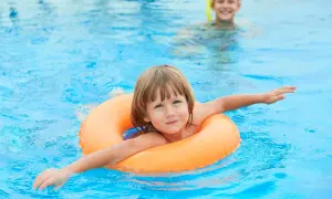 A happy child floats in an orange ring in a pool. Another child in goggles is in the background. This image reminds us when to stop swim lessons: when they're confident and enjoying the water!