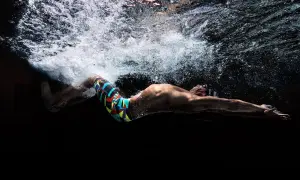 Underwater shot of a swimmer in a colorful tech suit, arms extended, with bubbles and light above.