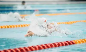 A swimmer in a white cap and goggles powers through the water, creating a splash. Their focused swimmer posture is evident as they propel forward in the lane.