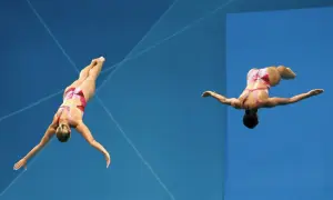 Two divers perform synchronized dives, showcasing their mastery of olympic diving terms as they execute a complex maneuver against a vibrant blue background.