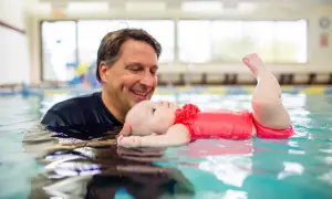 A father smiles as he holds his baby in a pool, showing that kids can learn to swim from a very young age.