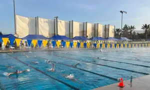 Swimmers race in lanes during a circle swim event. Blue and yellow banners line the pool, with tents and spectators in the background.