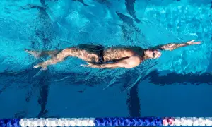 A male backstroke swimmer glides through the bright blue water of a pool, his body streamlined and arms extended.