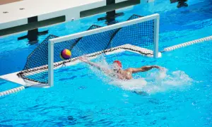 A water polo player in a red cap lunges for the ball near the net, splashing water. Intense underwater games are a thrilling spectacle.