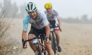 Smiles and determination on the faces of cyclists during a gravel triathlon. The lead rider, wearing a light blue and white jersey, grins as he navigates the dusty course.