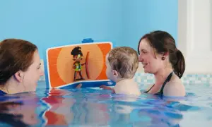 A toddler enjoys a fun toddler swim school lesson, looking at a colorful book with two smiling instructors in the pool.