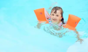 A happy young girl with bright blue eyes is floating in the pool, wearing orange arm floaties and a colorful swimsuit.
