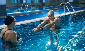 An adult swim coach instructs a swimmer in a pool, demonstrating proper arm stroke technique.