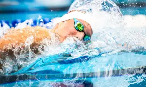 A swimmer's head emerges from the water, goggles reflecting the blue pool. Water splashes around them, a dynamic scene of fun swim drills.