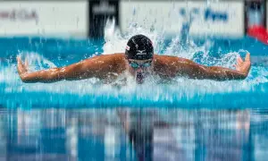 A swimmer in a black cap and goggles performs the butterfly stroke, arms extended, creating a splash as they take a breath.