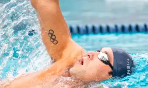 Swimmer in backstroke position, with Olympic rings tattoo on arm, mid-stroke, water splashing.