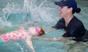 A child in a watermelon swimsuit and pink goggles floats on their back while a smiling instructor helps them practice treading water.