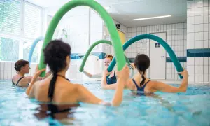 Women enjoy an aqua zumba class, holding colorful pool noodles for resistance and fun in the water.
