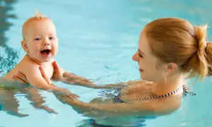 A happy baby laughs during baby and me swim classes, held by their mother in a bright blue pool.