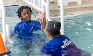 A young boy with dreadlocks in a blue rash guard gives a high-five to a swim instructor. This is a moment of connection during an autism swim lesson.