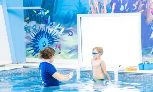 A swim instructor guides a young boy in goggles during 6 month swim lessons. The pool has a fun underwater mural.