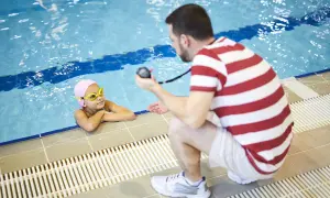 A mobile swim instructor crouches by the pool, holding a stopwatch, as a young girl in a pink cap and goggles rests her arms on the edge.