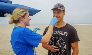 A woman in a blue shirt and gloves gently swabs a man's nose on a beach. He's wearing a cap and a t-shirt that reads "Trestles." This is part of a study to see if you can swim with a staph infection.