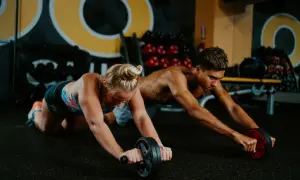 Two athletes, like swimmers training, perform ab roller exercises in a gym.