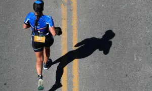A runner in blue triathlon apparel, bib number 107, runs on a road, casting a long shadow.