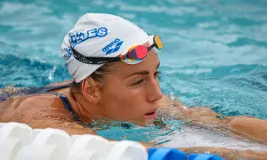 A female swimmer in a white cap and colorful goggles focuses intensely, her face emerging from the water during swim kick exercises.